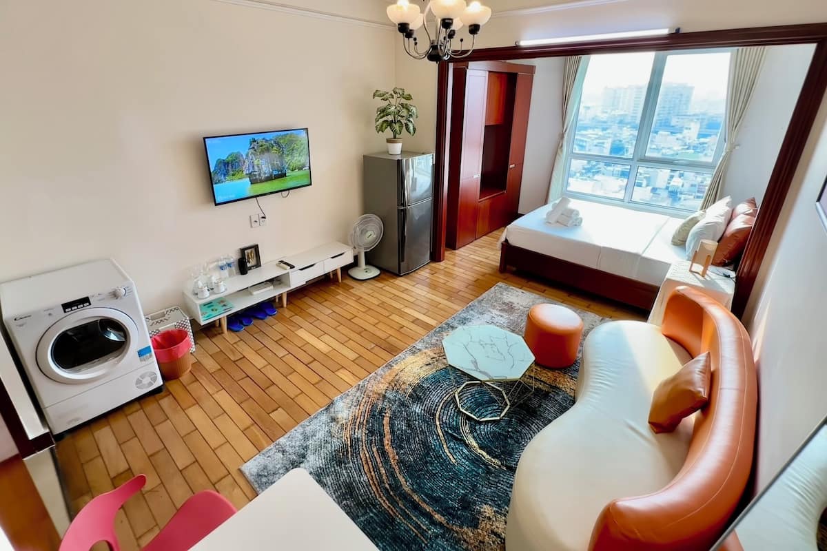 A modern hotel room with a large window, a washing machine, a flat-screen TV, a sofa, and a dining area.