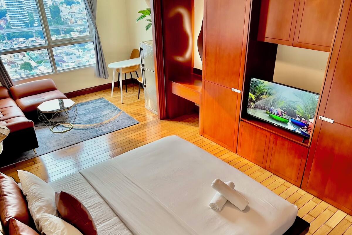 A modern hotel room with a large bed, a TV, a sofa, and a view of the city through large windows.