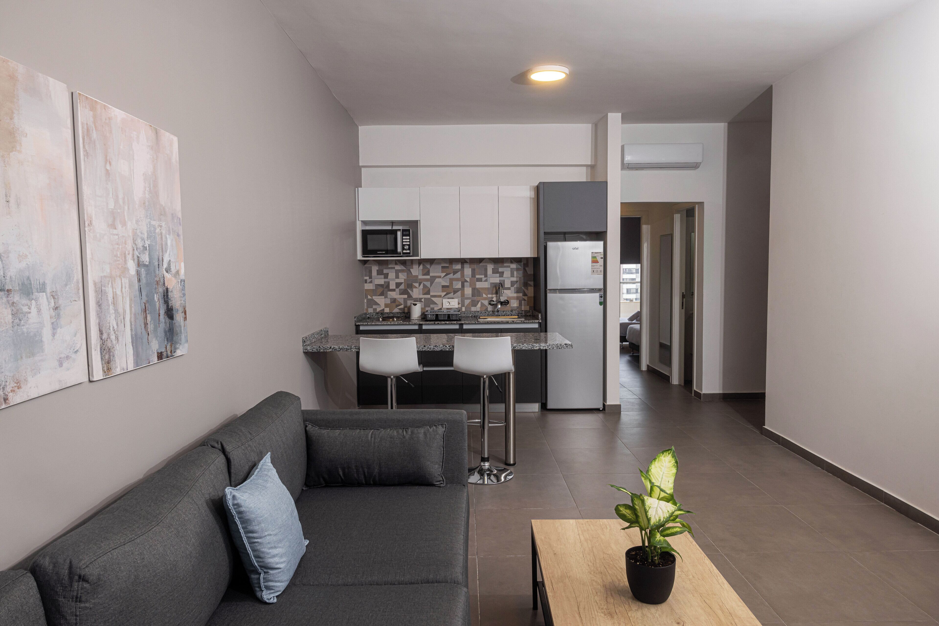 A modern apartment with a grey sofa, a small table with a plant, a kitchen with white cabinets and a refrigerator, and a hallway leading to another room.