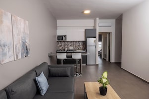Un apartamento moderno con un sofá gris, una mesita con una planta, una cocina con armarios blancos y un refrigerador, y un pasillo que conduce a otra habitación.