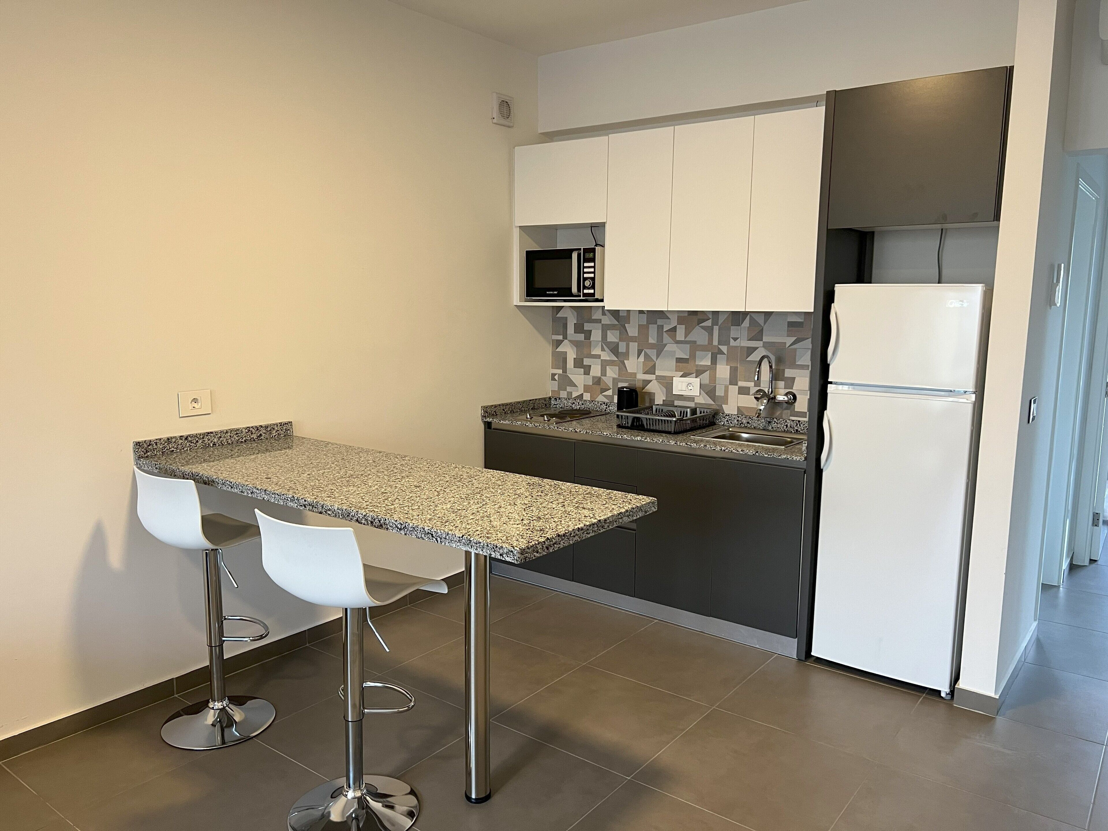 A compact kitchen with a granite countertop, two white bar stools, a microwave, a refrigerator, and a tiled backsplash.