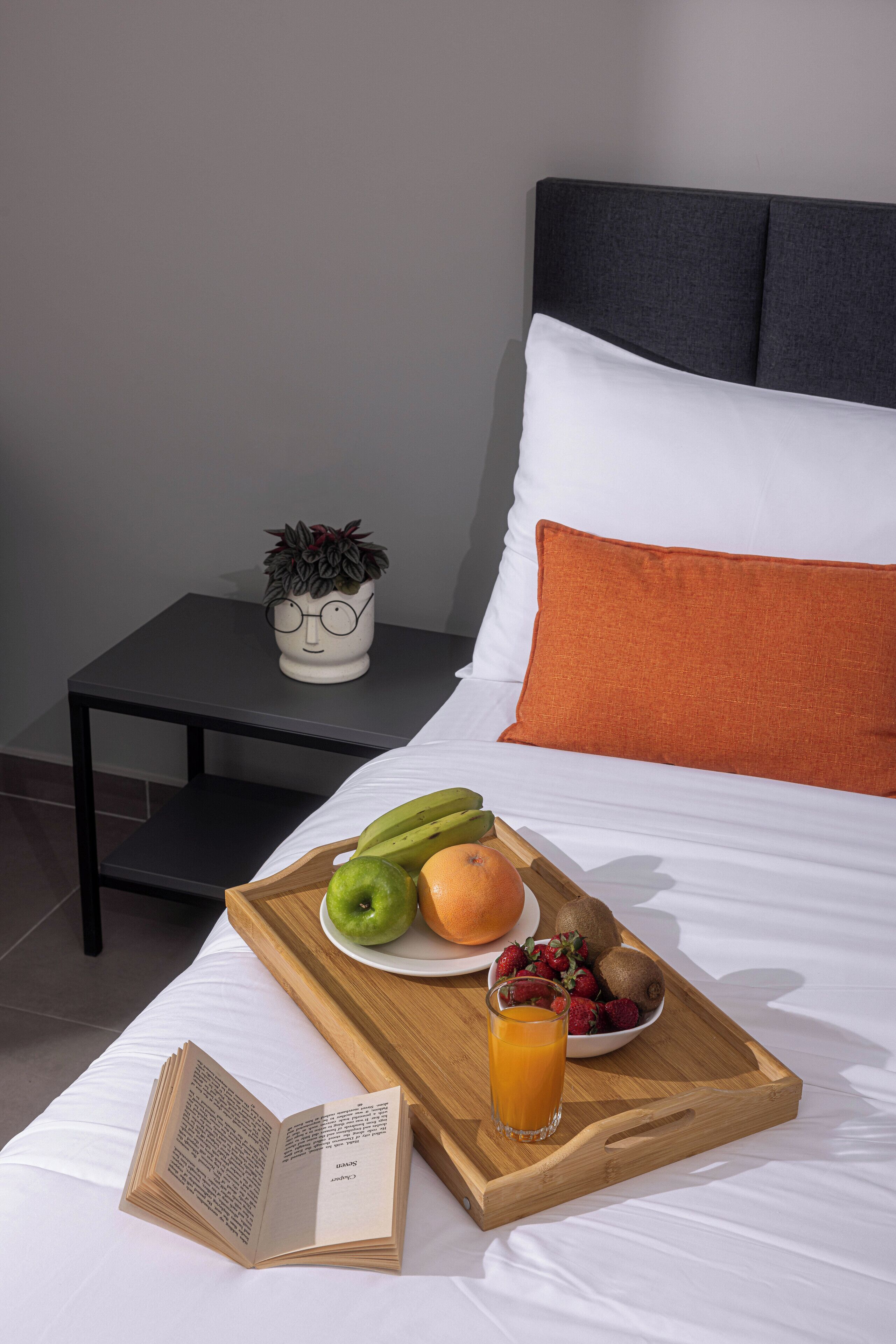 A neatly made bed with a tray of fruits and a glass of orange juice, an open book, and a small potted plant on a nightstand.