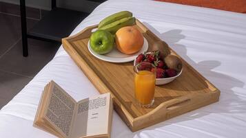 A neatly made bed with a tray of fruits and a glass of orange juice, an open book, and a small potted plant on a nightstand.