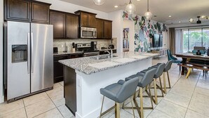 A modern kitchen with stainless steel appliances, a granite countertop island, bar stools, and pendant lighting. - Amazing Themed Villa with Private Pool (Kissimmee)
