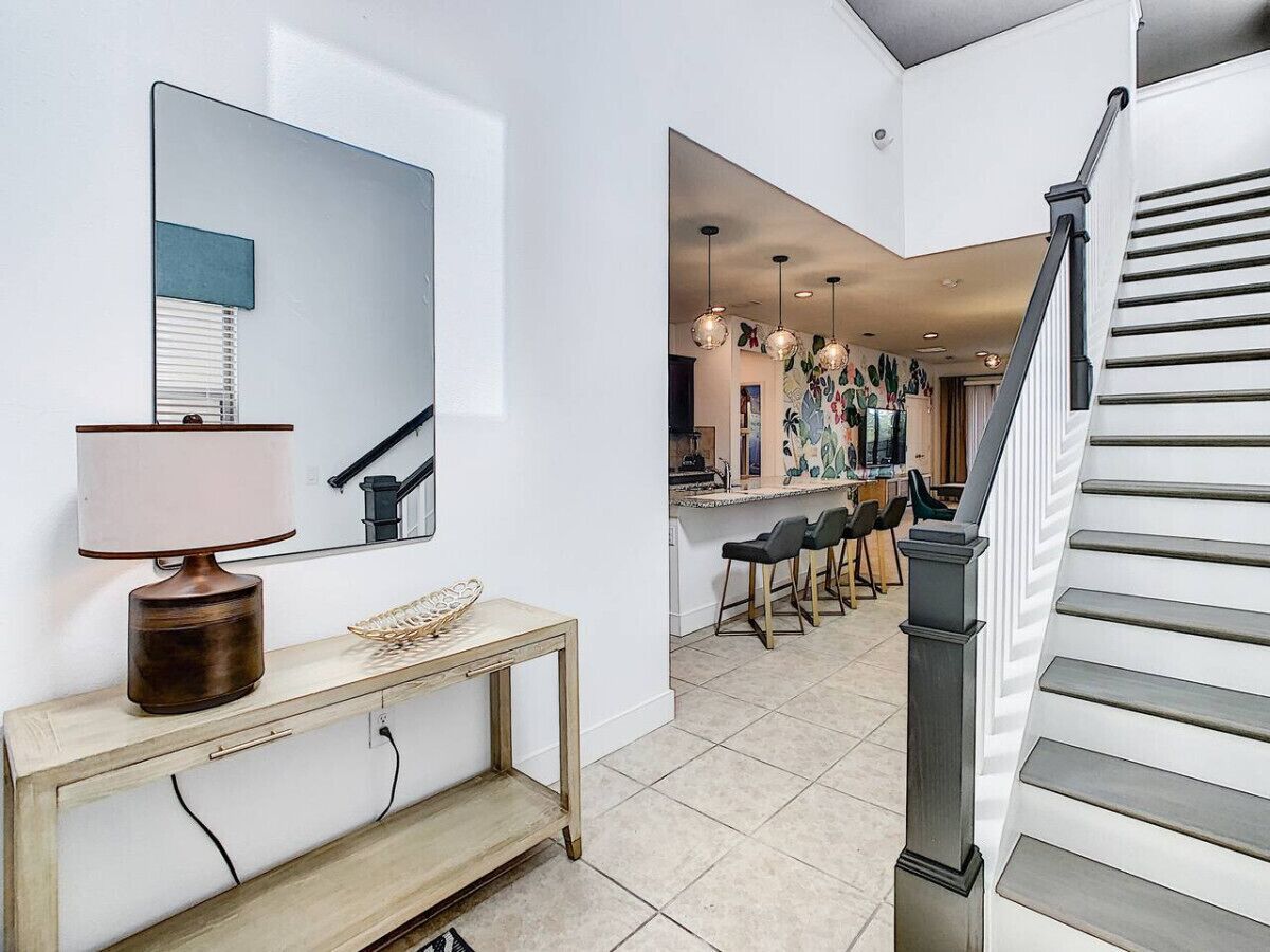 A modern interior with a wooden console table, a lamp, a mirror, and a staircase leading to a dining area with chairs and a kitchen in the background.