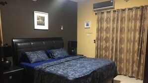 A bedroom with a dark brown headboard, a bed with a blue and white patterned bedspread, two bedside tables with lamps, and a large window with curtains. - Shortlet Apartment Milverton Estate (Lekki)