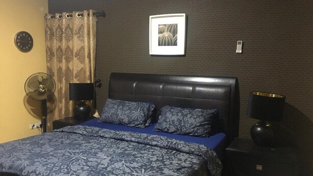 A bedroom with a dark leather headboard, a blue patterned bedspread, two matching pillows, a bedside table with a lamp, a wall clock, and a framed picture on the wall. - Shortlet Apartment Milverton Estate (Lekki)