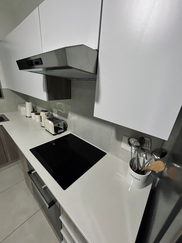 A modern kitchen with white cabinets, a black induction cooktop, a stainless steel range hood, and a utensil holder with a wooden spoon. - Ventura Estate  (Randburg)