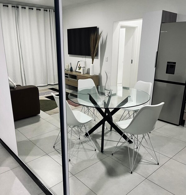 Een moderne keuken met een glazen eettafel, witte stoelen en een roestvrijstalen koelkast.