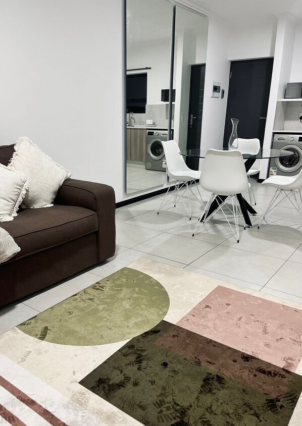 Ein modernes Wohnzimmer mit einem braunen Sofa, weißen Stühlen und einem Glastisch. Ein großer Spiegel spiegelt den Raum wider und im Hintergrund steht eine Waschmaschine.