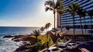 Un hôtel situé sur la côte, offrant une vue sur l’océan, des palmiers et un parking avec des voitures.