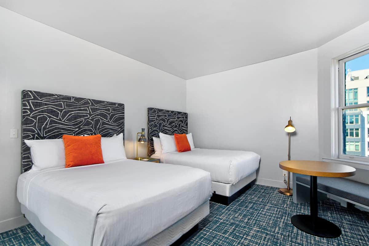 A hotel room with two beds, a small round table, a lamp, and a window with a city view.