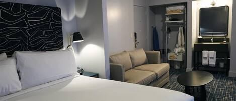 A hotel room with a bed, a sofa, a small table, a mirror, a wardrobe, and a shower area.