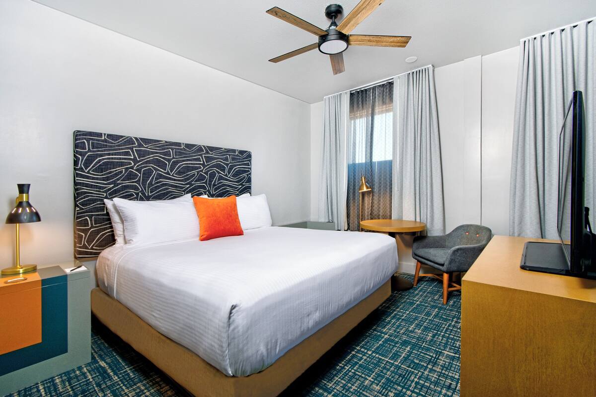 A hotel room with a bed, a desk, a chair, a ceiling fan, and a window with curtains.