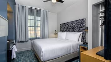 Une chambre d’hôtel avec un lit, un ventilateur de plafond, une fenêtre avec des rideaux, une tête de lit à motifs, une table de chevet et un bureau en bois.