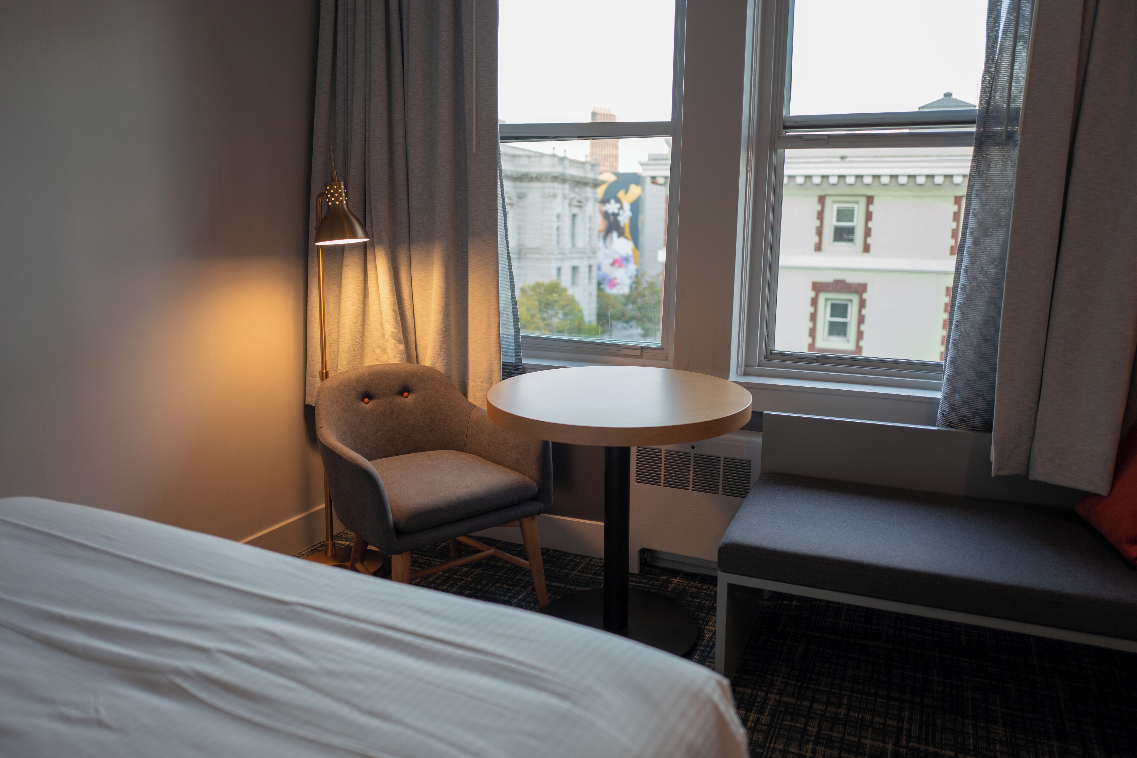 A hotel room with a bed, a small round table, a chair, and a window with a view of buildings.