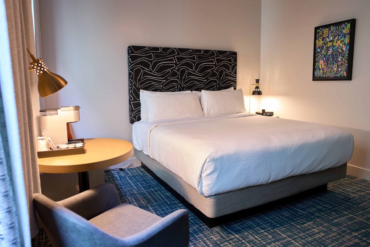 A hotel room with a bed, a desk with a lamp, a chair, and a wall art piece.