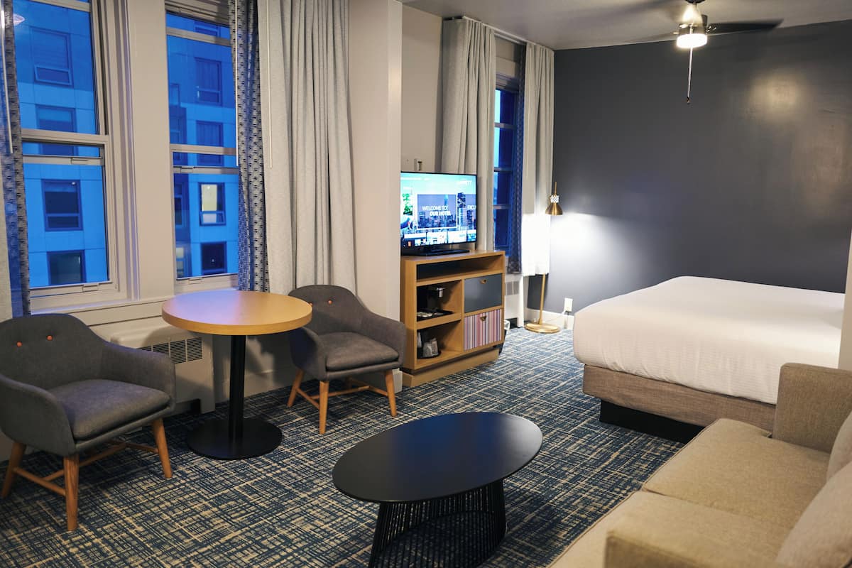 A hotel room with a bed, two armchairs, a small table, a TV on a stand, and a bookshelf.