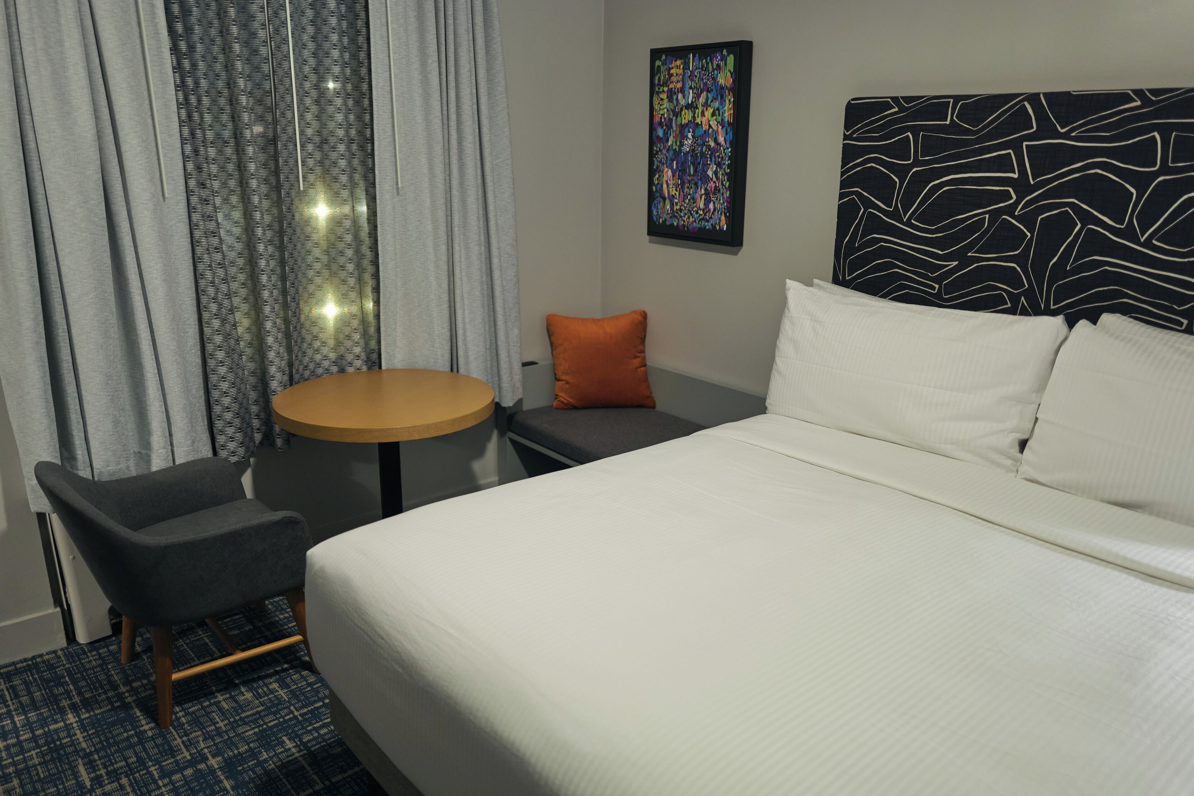 Une chambre d’hôtel avec un lit, une table ronde, une chaise, un petit coussin orange et une tête de lit à motifs.