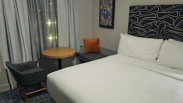Une chambre d’hôtel avec un lit, une table ronde, une chaise, un petit coussin orange et une tête de lit à motifs.