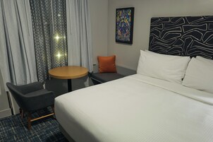 A hotel room with a bed, a round table, a chair, a small orange pillow, and a patterned headboard.