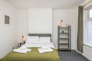 Quarto de hotel bem arrumado, com uma cama, mesa de cabeceira com abajur, prateleira com objetos decorativos e uma janela com cortinas.