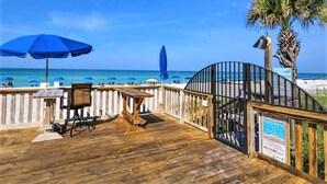 Une terrasse en bois offrant une vue sur la plage, avec une table, des chaises et plusieurs parasols bleus.