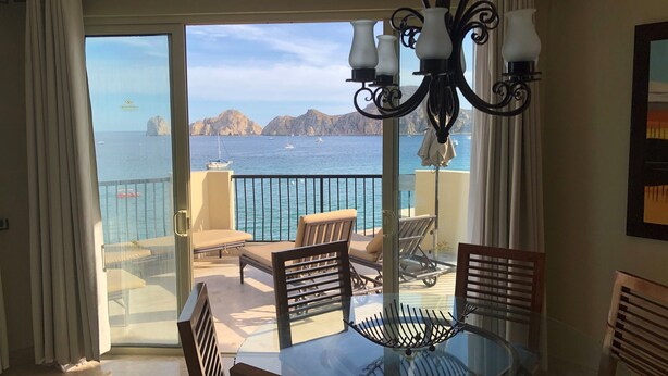 A balcony with a glass table and chairs, overlooking a sea view with boats and rocky terrain.