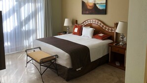 A hotel room with a large bed, two bedside tables with lamps, a chair, a ceiling fan, a painting on the wall, and a window with curtains.
