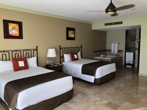 Habitación de hotel con dos camas, una mesita de noche, una lámpara, un ventilador de techo y un baño visible a través de una puerta abierta.