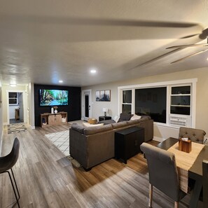 A modern living room with a sectional sofa, a dining area with a table and chairs, and a large TV mounted on the wall.