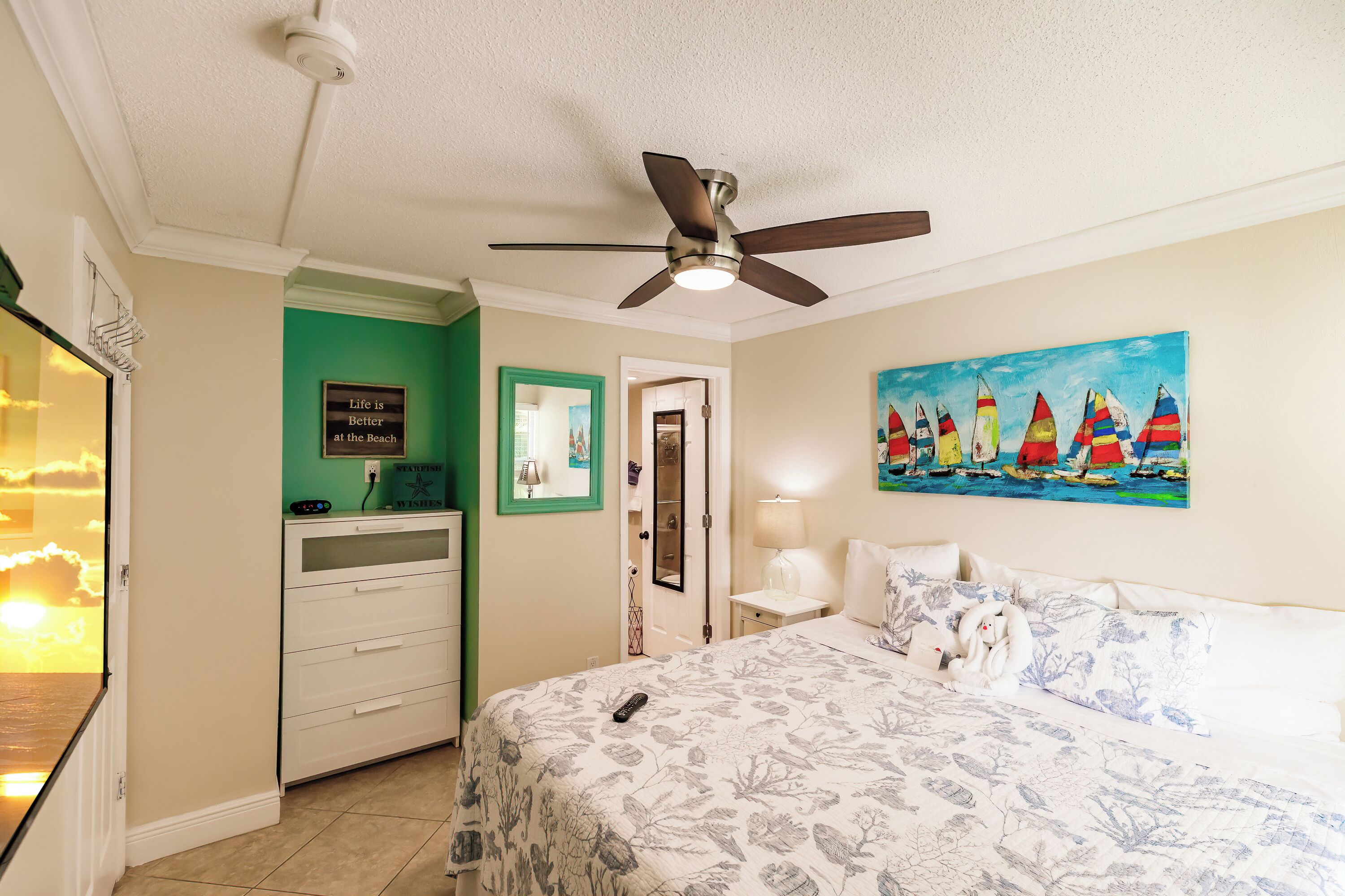 A bedroom with a bed, a ceiling fan, a painting of sailboats, a dresser, and a door leading to another room.