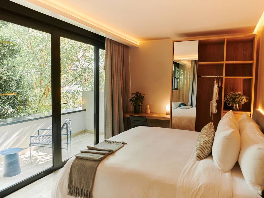 Habitación de hotel moderna con una cama grande, un escritorio, un espejo y un balcón con vistas a árboles.