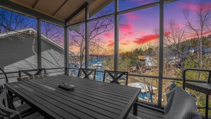 Un espacio de estar techado al aire libre con una mesa y sillas, con vista a un barrio residencial y una masa de agua durante la puesta de sol.