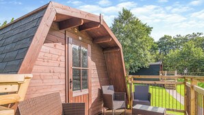 A wooden cabin with a sloped roof, a deck with wicker furniture, and a view of a green landscape.