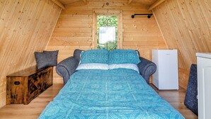 A cozy attic bedroom with a wooden sloped ceiling, a bed with patterned bedding, a small white refrigerator, and a bench with cushions.