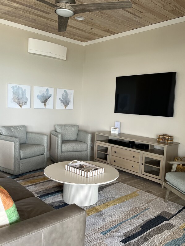Una sala de estar con ventilador de techo, un televisor de pantalla plana, dos sillones grises, una mesa de centro con revistas, un tapete estampado y un mueble de madera.