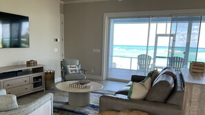 A spacious living room with a large window overlooking the beach, a comfortable sofa, a coffee table, and a TV mounted on the wall.