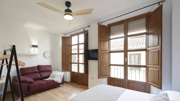 Een slaapkamer met een bed, een rode bank, houten luiken, een plafondventilator en een televisie.