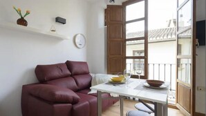 Een woonkamer met een rode bank, een klein tafeltje met een schaal en wijnglazen, een klok aan de muur, en een balkon met een hekwerk.