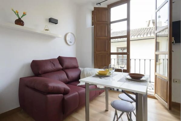 Un salon avec un canapé rouge, une petite table sur laquelle se trouve un saladier et des verres à vin, une horloge au mur et un balcon avec une balustrade.