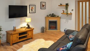 Een moderne woonkamer met een zwarte leren bank, een houten entertainmentkast, een flatscreen-tv, een open haard en wandplanken met decoratieve objecten.