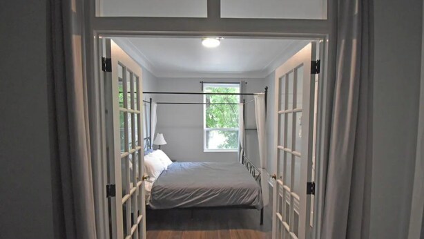 Een smalle gang die leidt naar een slaapkamer met een bed, een raam en een deur.