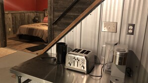 Une cuisine compacte équipée d’un grille-pain, d’une machine à café et d’un petit réfrigérateur. On aperçoit un lit avec un mur rouge et un lambris en bois à l’arrière-plan.