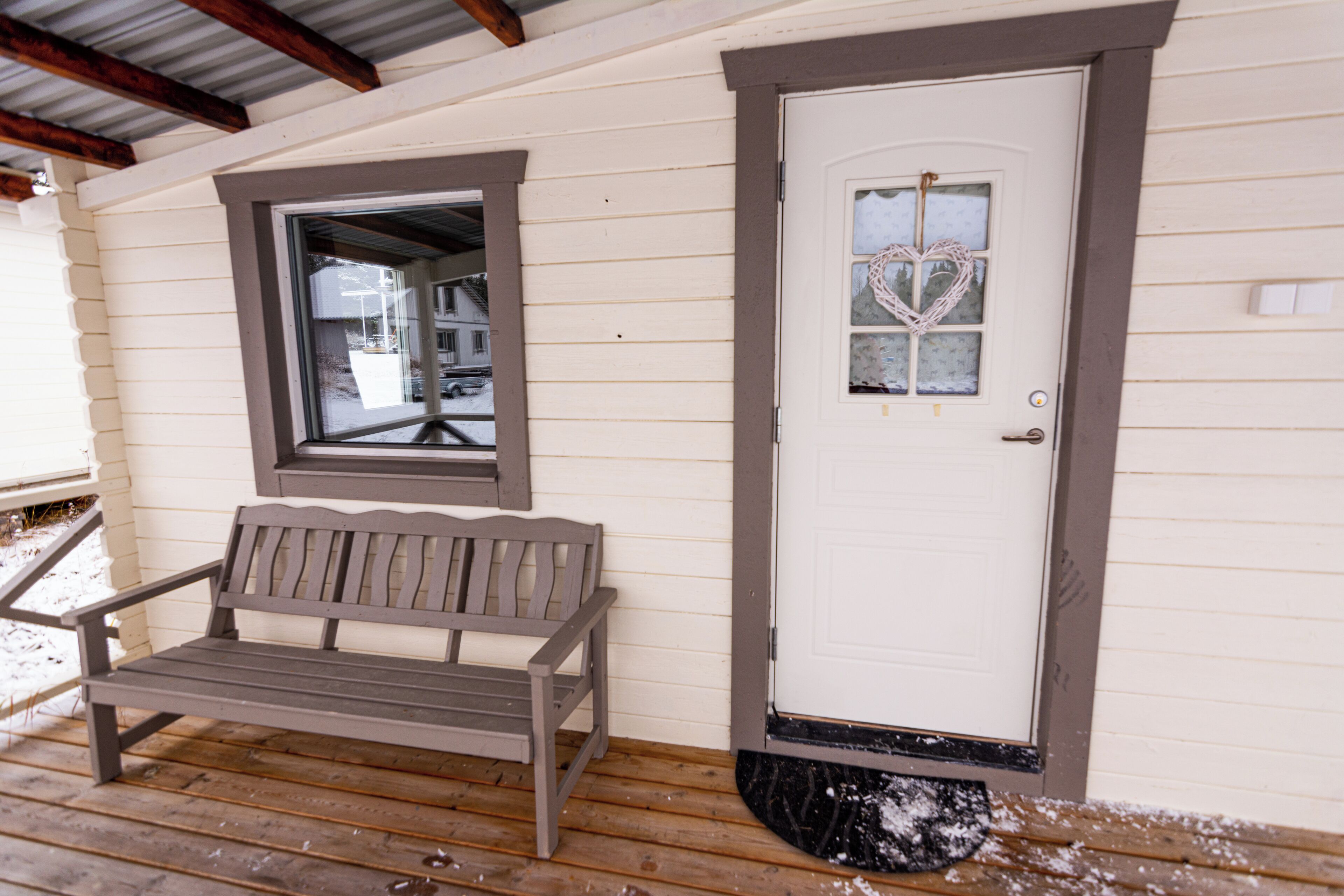 A wooden bench on a porch with a white door and a heart-shaped window decoration.
