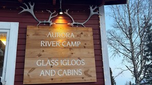 Un letrero de madera con el texto 'Aurora River Camp Glass Igloos and Cabins' sujeto a un edificio de fachada roja.