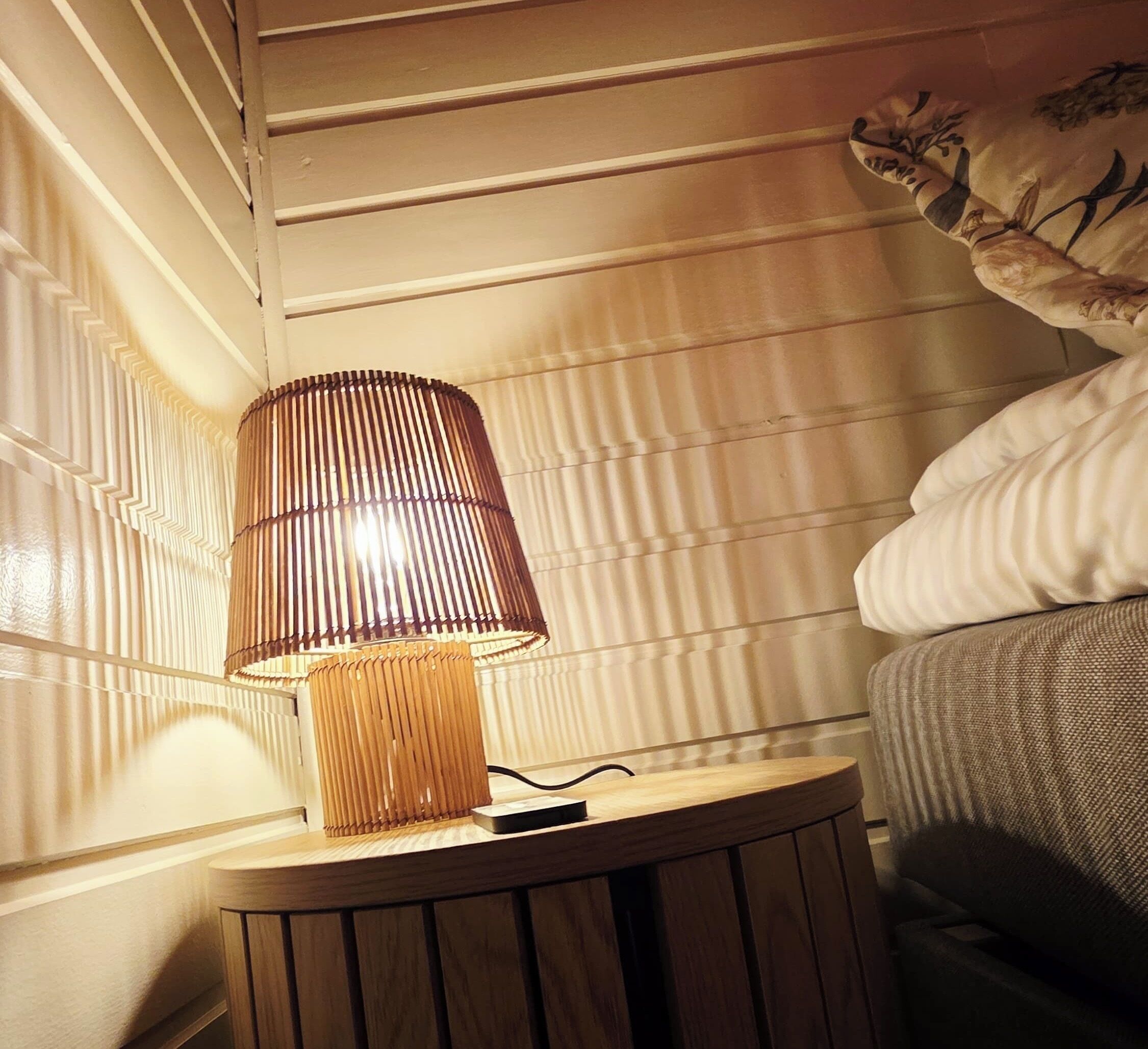 A bedside table with a lamp, a bed, and a wall with vertical lines.