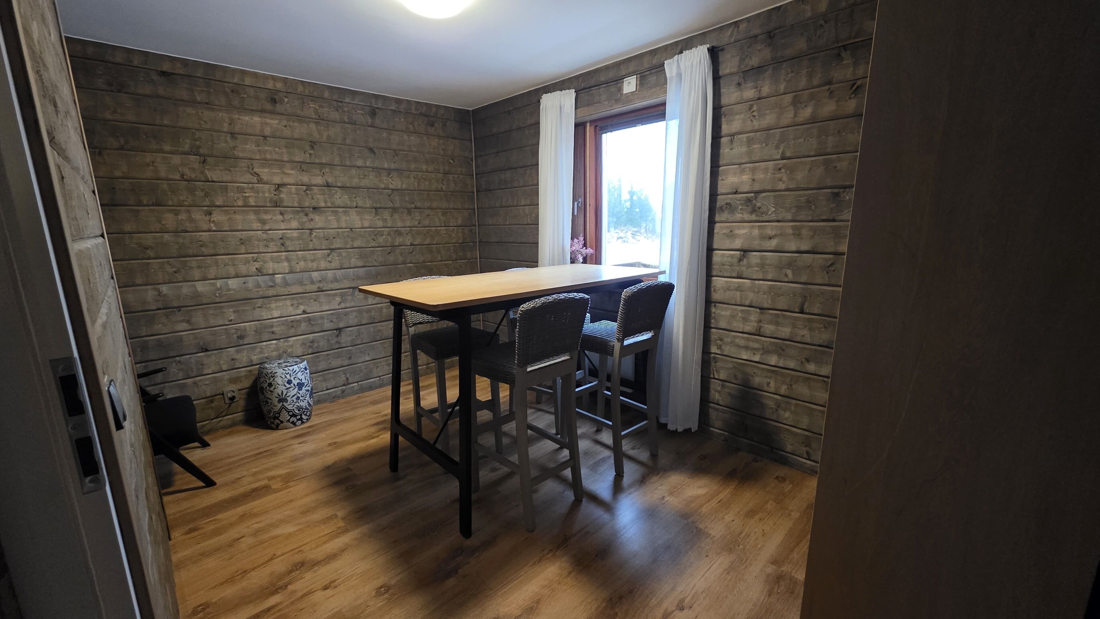 A room with a wooden floor, a wooden table with four chairs, a window with white curtains, and a small decorative vase.