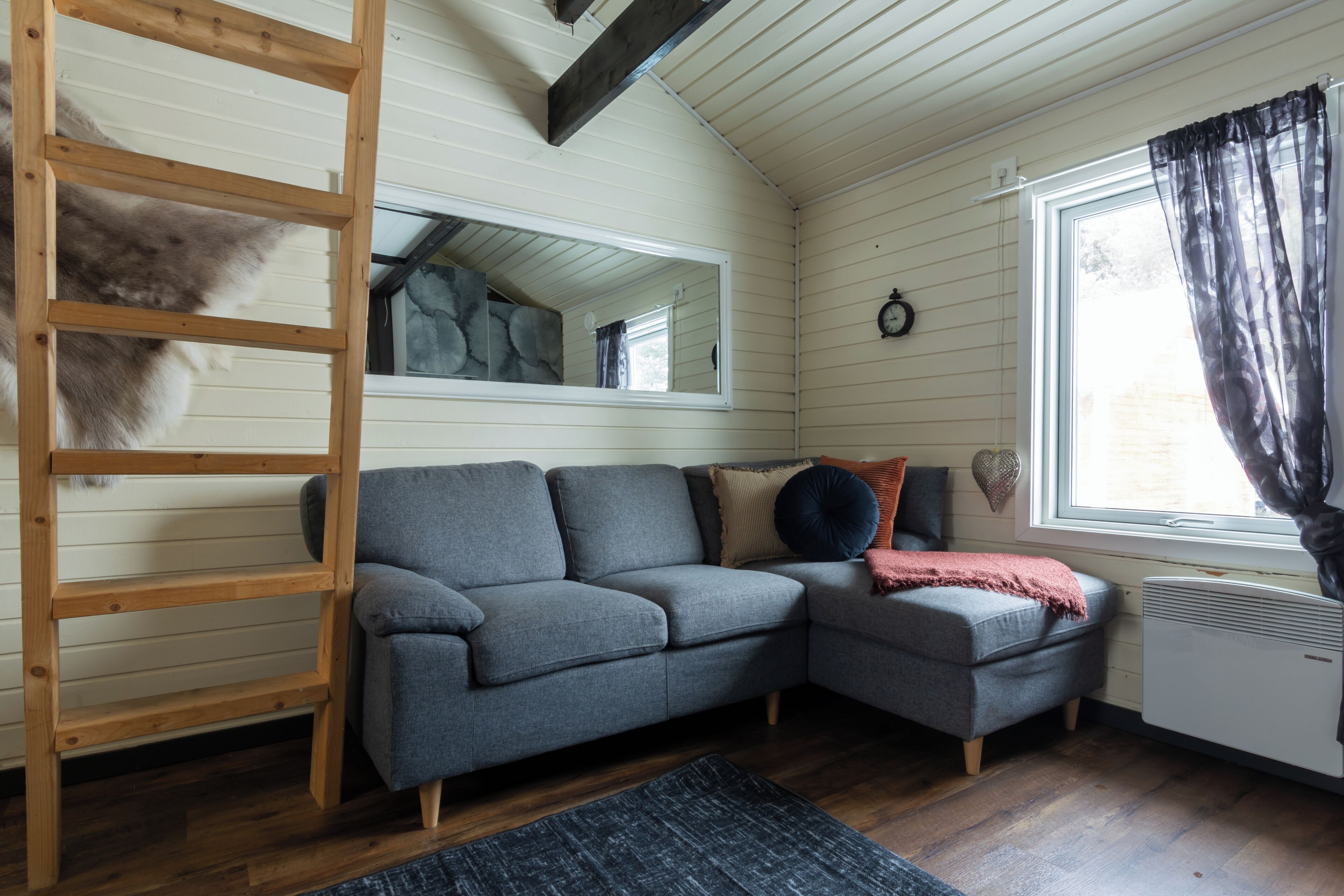A cozy room with a gray sectional sofa, a wooden ladder, a large mirror, and a window with curtains.