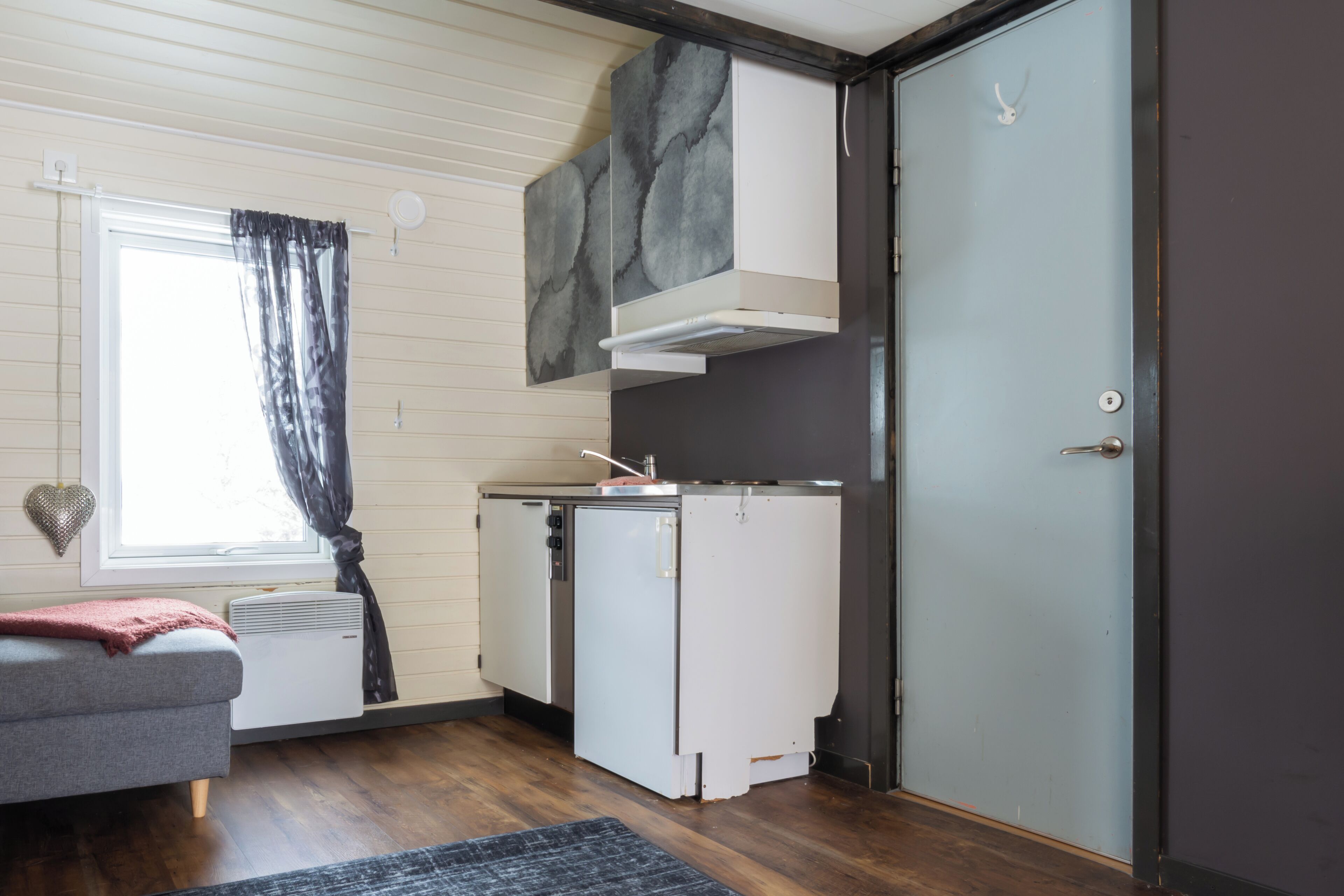 Una habitación pequeña con cama, kitchenette, ventana con cortinas y puerta.